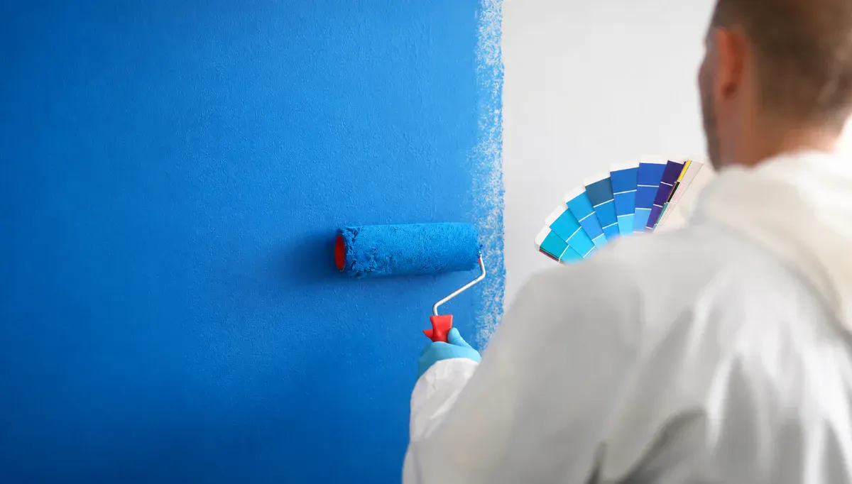Peintre en bâtiment appliquant au rouleau une peinture bleue sur un mur intérieur, nuancier à la main